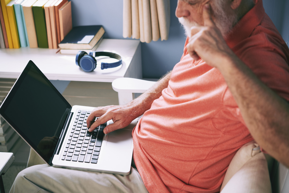 Older adult in a coral shirt using a laptop at home with books and headphones nearby.